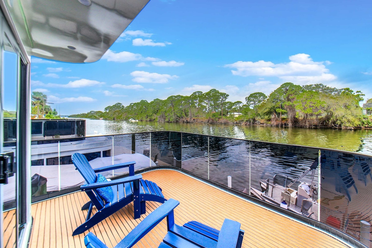 A spacious deck features blue Adirondack chairs arranged for relaxation, overlooking calm waters. Lush greenery is seen along the shoreline while boats are moored nearby. Glass railings provide unobstructed views, creating a serene atmosphere for enjoying the natural surroundings.