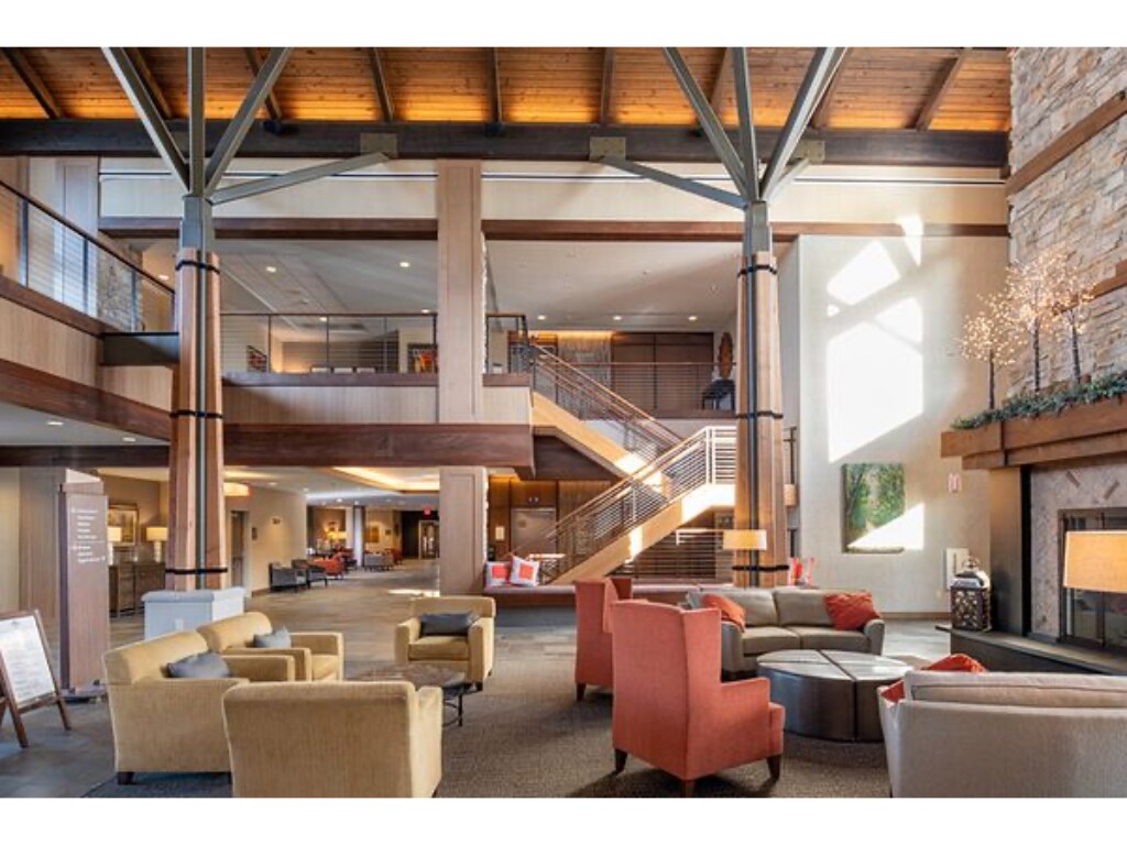 The spacious lobby is characterized by high ceilings and natural light streaming through large windows. A cozy seating area features a circular coffee table surrounded by various armchairs. Wooden beams and a stone fireplace add to the warm ambiance of the common space.