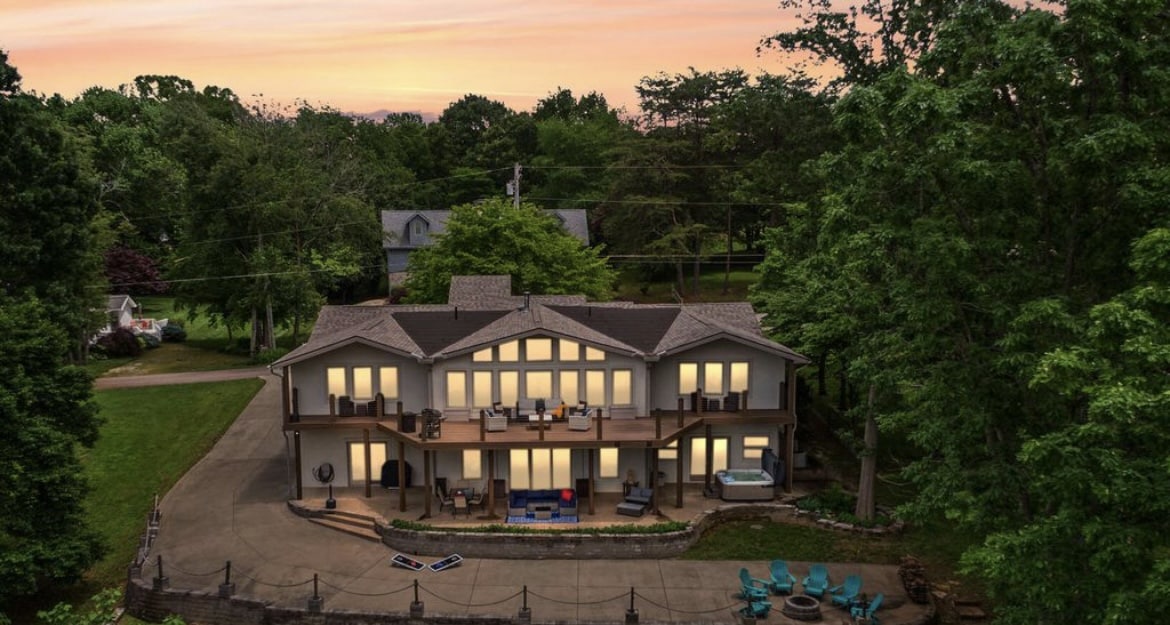 The villa is showcased from an elevated perspective during sunset, highlighting its spacious two-story exterior and large windows reflecting the warm evening light. The surrounding greenery enhances the serene atmosphere, while outdoor seating arrangements are visible on the expansive deck.