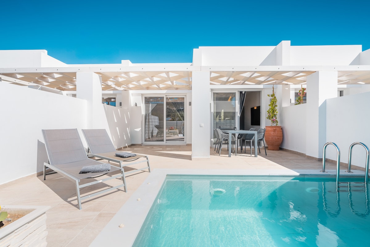 A private outdoor area features a soothing pool and two lounging chairs positioned nearby. An open terrace is visible alongside the pool, with a dining table set for four. Sunlight brightens the minimalist design, enhancing the relaxing ambiance of this space.