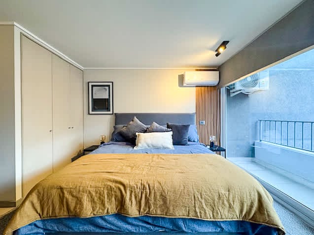 A spacious bedroom features a comfortable bed dressed in layered bedding, complemented by decorative pillows. Soft lighting illuminates the room, while large windows provide outdoor views and natural light. A minimalist closet is positioned to one side, maintaining a clean, uncluttered space.