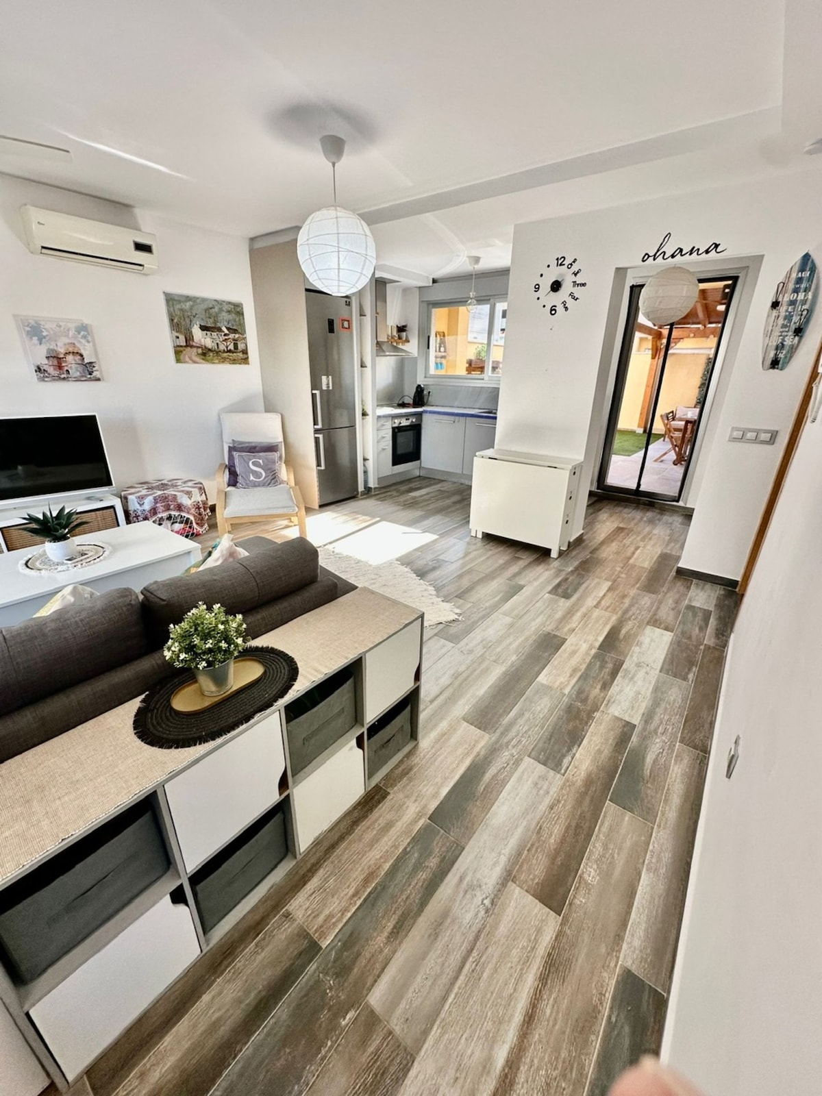 A modern open-plan living area is featured, showcasing a comfortable sofa with decorative cushions. A large TV sits on a media console. The kitchen, equipped with stainless steel appliances, is adjacent. Bright light streams through windows, highlighting the warm wooden flooring and welcoming atmosphere.