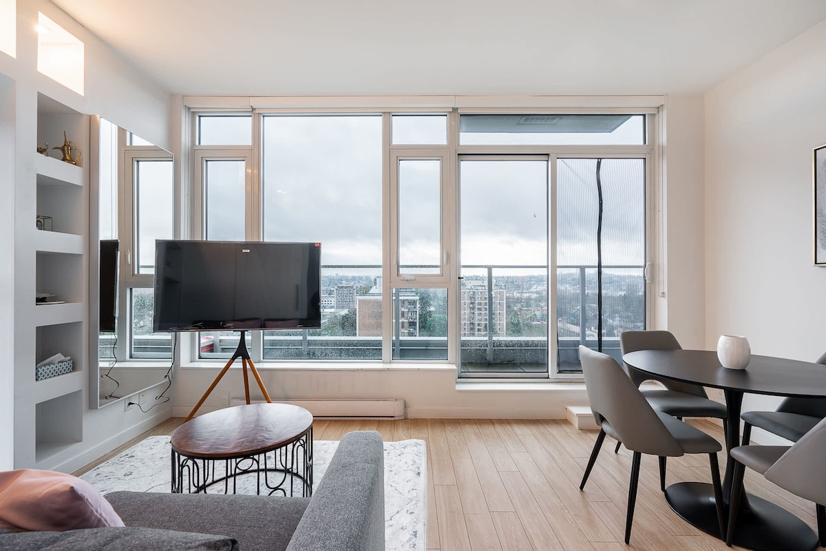 An open-concept living area is featured, showcasing large floor-to-ceiling windows that allow for expansive city views. A comfortable gray sofa is positioned adjacent to a round dining table with chairs. A flat-screen television stands on a modern stand, enhancing the contemporary feel of the space.