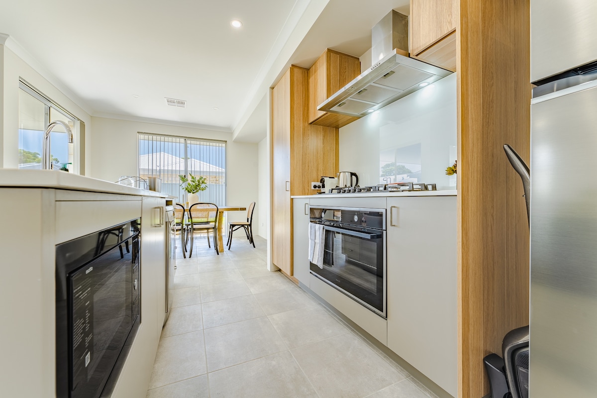 The kitchen is equipped with modern appliances, including a gas stovetop and an electric oven. An adjoining dining area features a table with seating for six, illuminated by natural light from nearby windows. The cabinetry is crafted from light wood, contributing to a bright ambiance.
