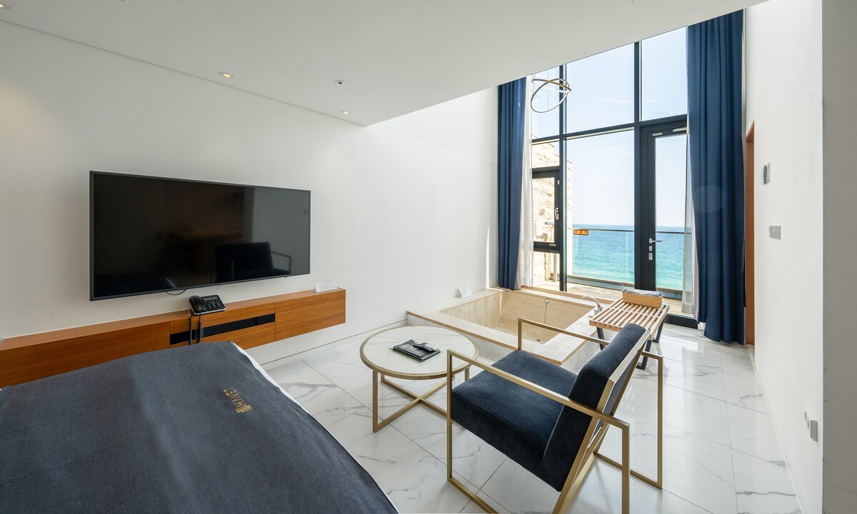 A modern living area features a flat-screen television mounted on a warm wooden console. A stylish armchair and a coffee table are arranged beside a large window, offering a view of the sea. Soft natural light fills the space, enhancing its open feel.