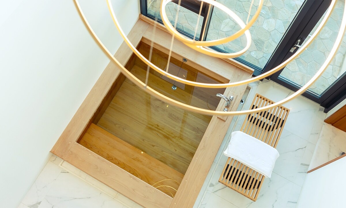 An overhead view of a wooden hot tub is captured, situated beneath a modern light fixture. Natural light floods the area, highlighting the spa's wooden accents and the marble flooring beside a wooden bench with a folded towel.