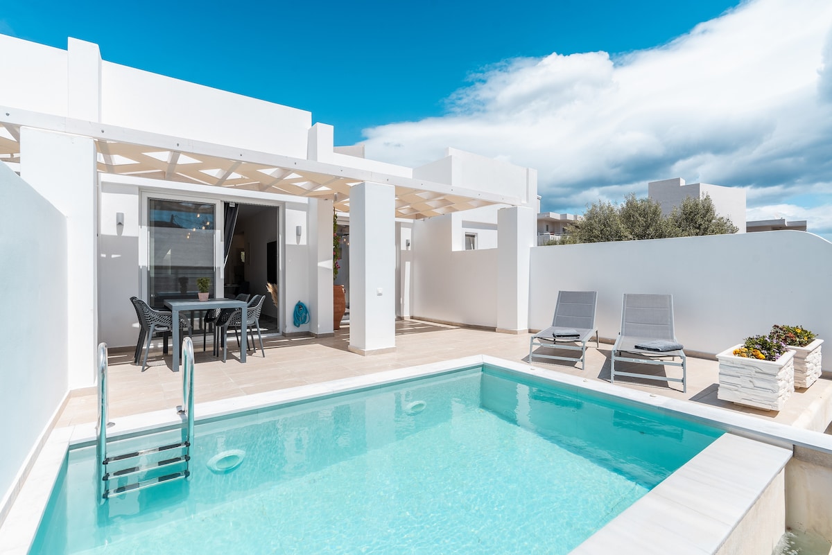 An outdoor area is presented, featuring a private pool with clear water. Lounge chairs are positioned nearby, and a shaded dining table with chairs offers space for al fresco meals. White walls create a clean aesthetic, with blue skies visible above.