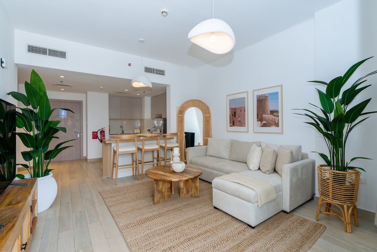 The living area is designed with natural elements featuring a large, light-colored sectional sofa accompanied by a wooden coffee table. A textured rug covers the floor, and potted plants are placed strategically to enhance the space. Two framed images adorn the wall, contributing to a modern aesthetic.
