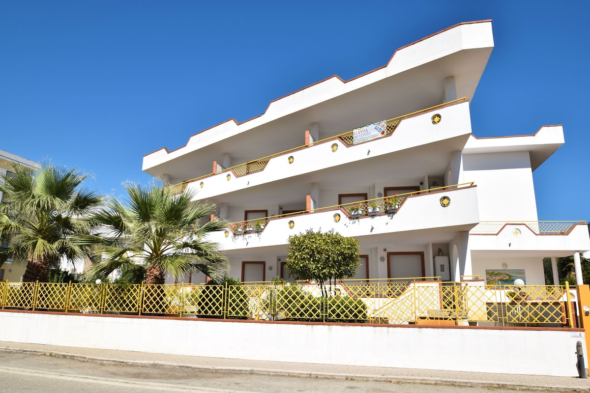 The modern multi-level building is showcased against a clear blue sky. A balcony adorned with plants and a vibrant yellow railing adds character. Palm trees and manicured greenery frame the entrance, creating a welcoming aspect for guests.