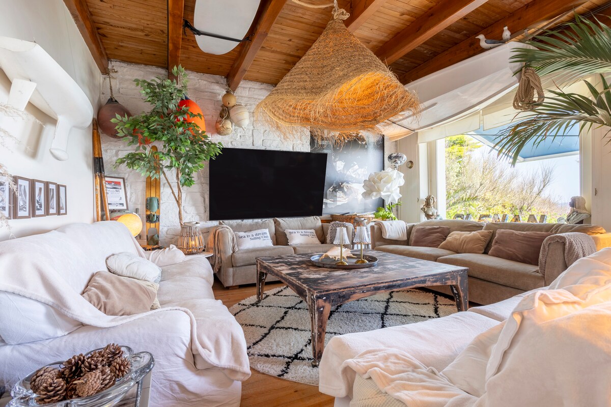 A spacious living area is characterized by a high wooden ceiling and large windows offering views of the outdoors. A large flat-screen TV is mounted on the wall, while comfortable sofas surround a central coffee table adorned with decorative items. A large woven light fixture hangs above, and greenery adds a touch of nature.