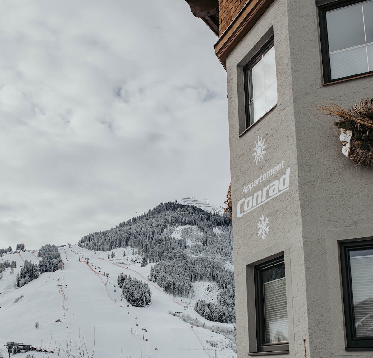 The image captures a winter landscape featuring a ski resort in the background, with snow-covered slopes and trees. The building, bearing the name 'Appartement Conrad,' is situated nearby, showcasing a blend of natural beauty and cozy accommodation.