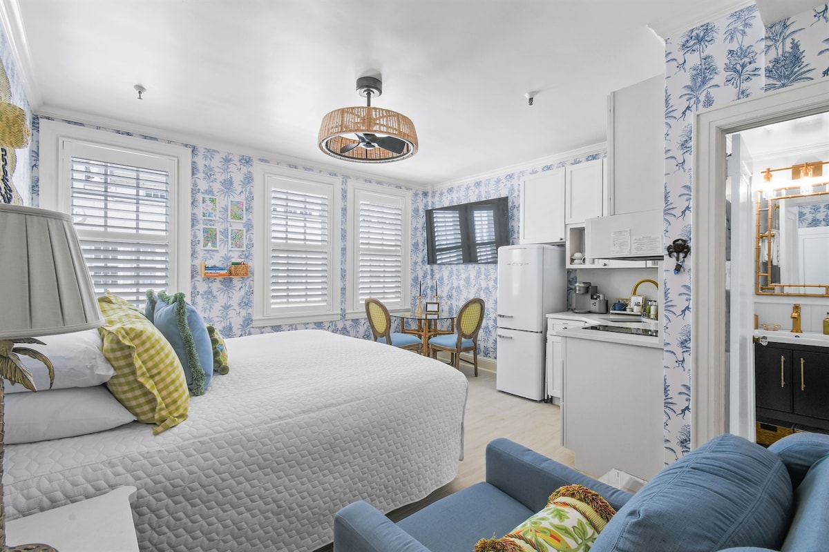 The room features bright blue wallpaper and ample natural light from multiple windows, complemented by a spacious bed adorned with colorful pillows. A small dining area with two chairs is visible next to a fully equipped kitchen, and a stylish bathroom is accessible through a door.