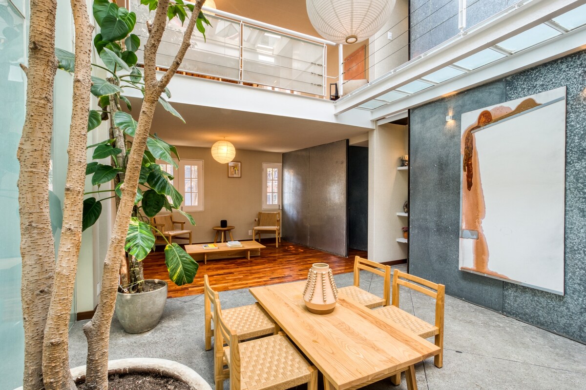 An open interior space is highlighted by a large, potted tree and a wooden dining table surrounded by woven chairs. Large windows provide natural light, while a light fixture hangs above. A large artwork adorns the wall, adding visual interest to the area.
