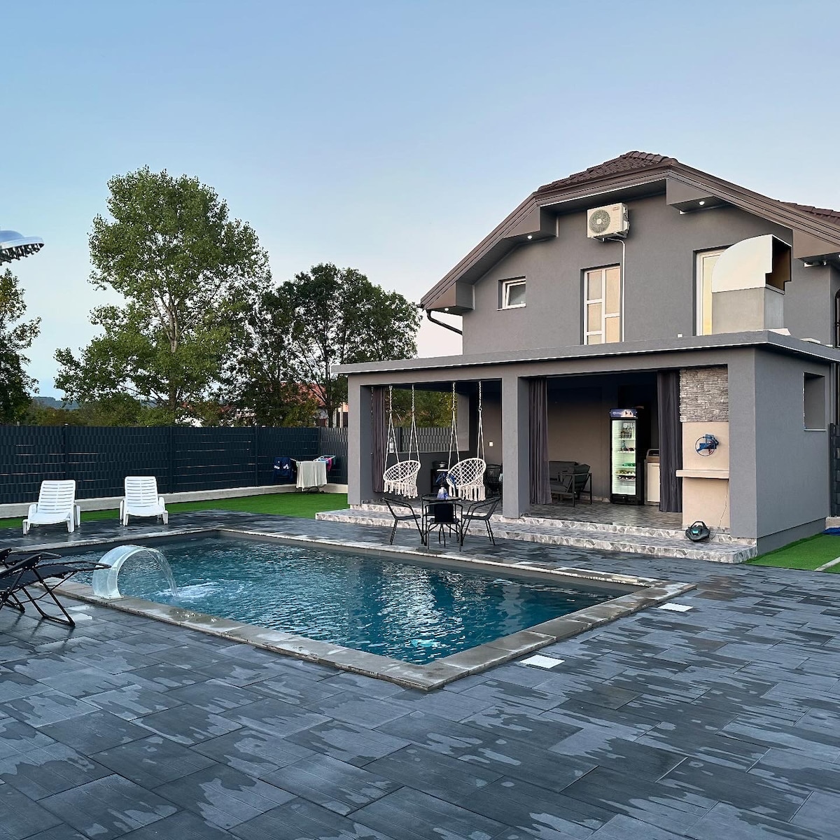 A modern house is showcased with a sleek outdoor pool and spacious patio area. Lounge chairs are arranged around the pool, providing a comfortable space for relaxation. The building features large glass doors that open to the serene surroundings, complemented by a well-maintained lawn.