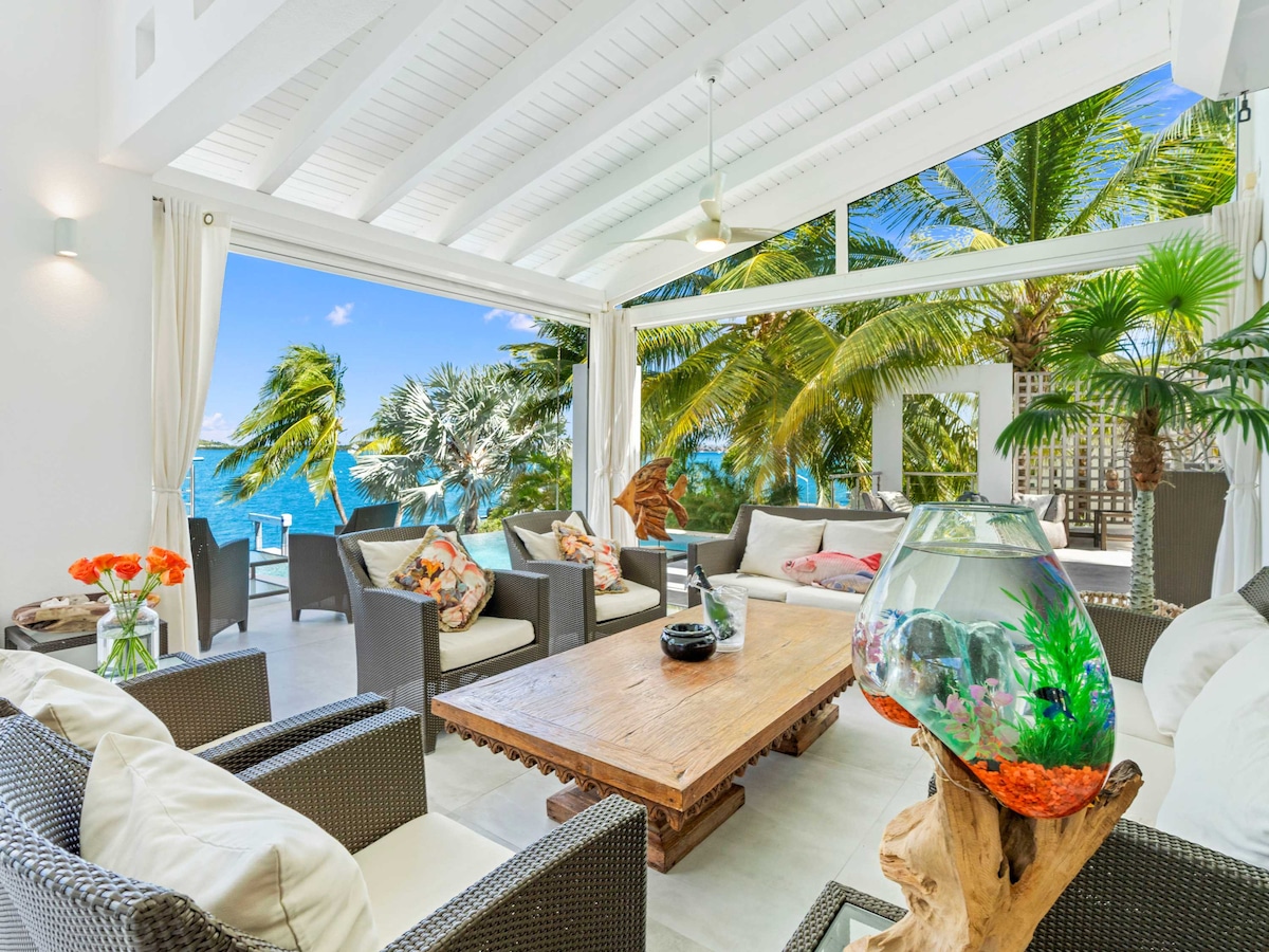 A spacious seating area is highlighted by natural light flowing through large glass doors. Comfortable wicker furniture surrounds a wooden coffee table. Tropical plants and a fishbowl are visible, framing views of the ocean and palm trees in the background.