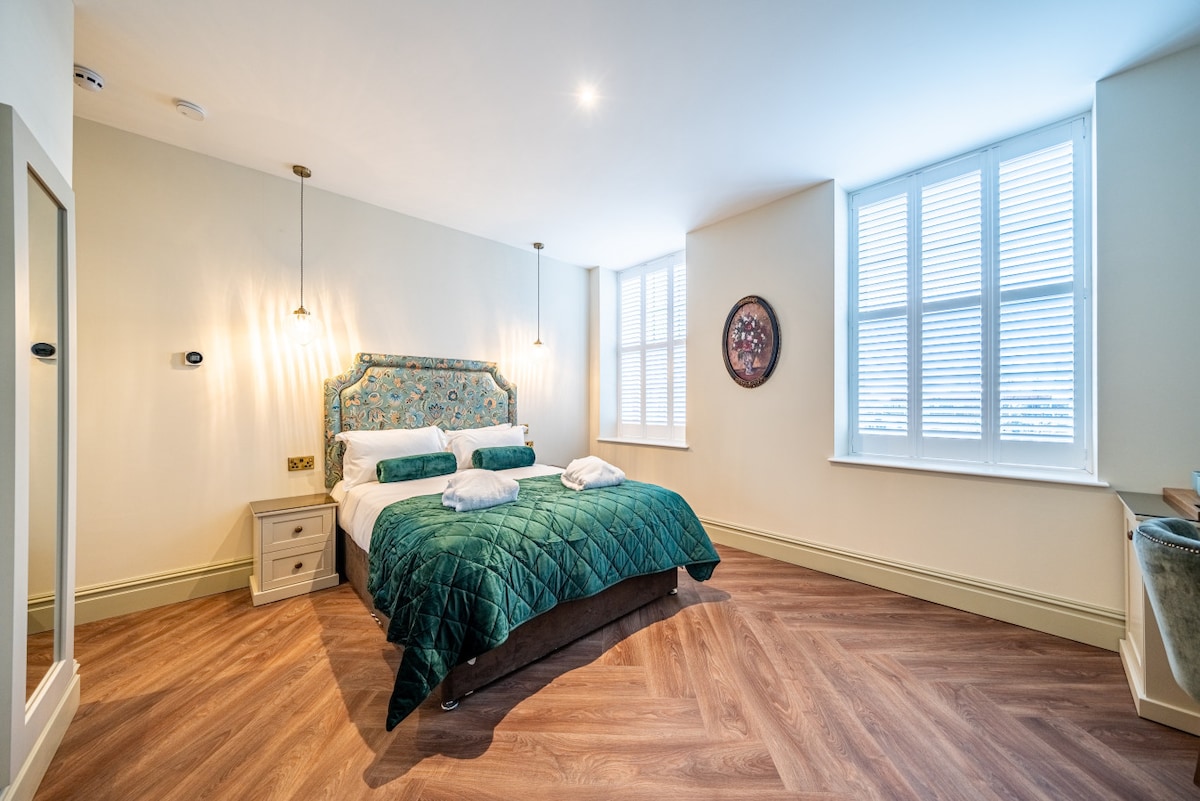 A spacious bedroom features a luxurious king-size bed with an elegant green quilt. Light-colored walls complement the wooden floor, while large sash windows with shutters allow natural light. A framed artwork hangs on the wall, and a stylish bedside table adds functionality.