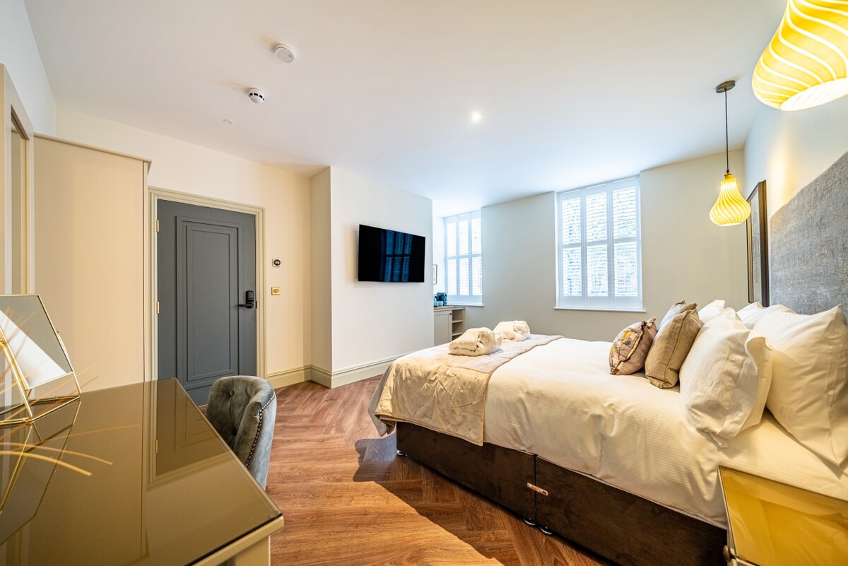 The spacious room features a king-size bed with decorative pillows. Natural light streams through sash windows with shutters. A flat-screen TV is mounted on the wall across from the bed. A stylish desk with a chair is positioned nearby, enhancing the room's functionality.