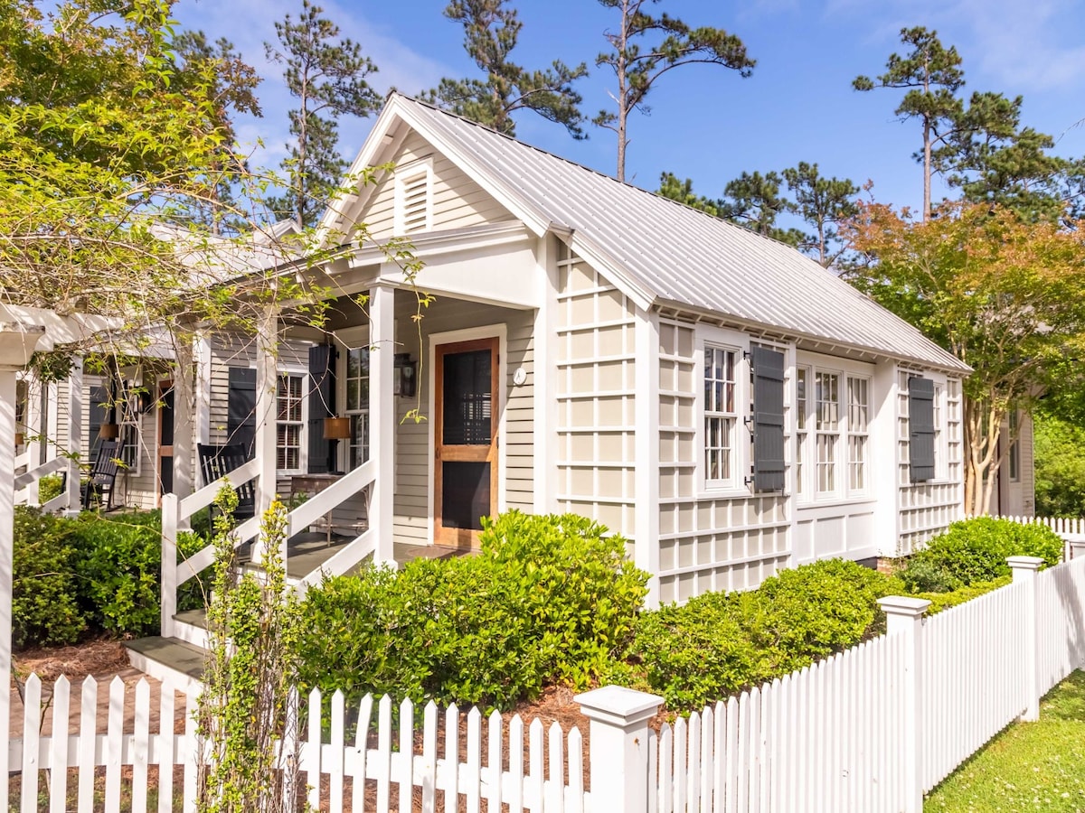 The exterior of the cottage is presented, featuring a white picket fence and lush hedges that frame the entrance. The house showcases a classic design with charming shutters and a covered porch, hinting at a welcoming ambiance. Tall trees provide a natural backdrop.
