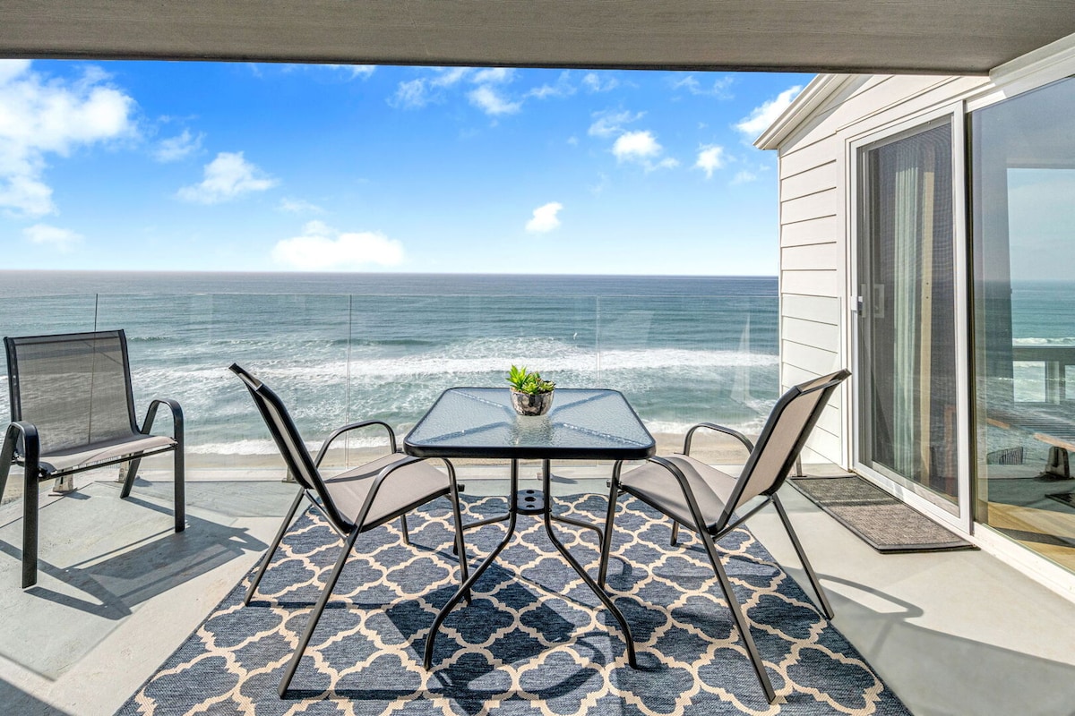 A private balcony offers an outdoor dining table surrounded by four chairs, with a patterned area rug beneath. The ocean view stretches into the distance, framed by a clear glass railing that accentuates the relaxing coastal atmosphere.