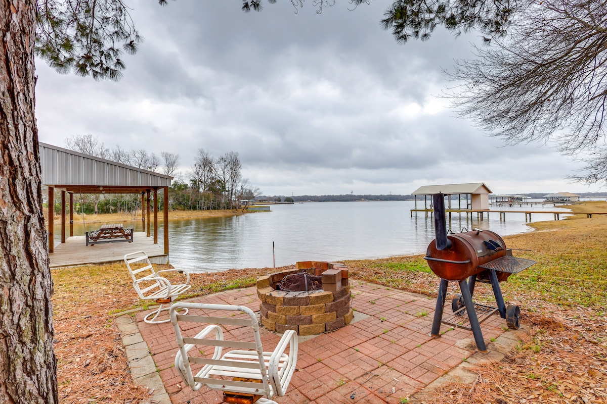 A welcoming outdoor area features a stone fire pit surrounded by several white chairs. In the background, a covered seating area is visible near the water's edge, alongside a charcoal grill and expansive lake views. Overcast skies reflect a peaceful ambiance.