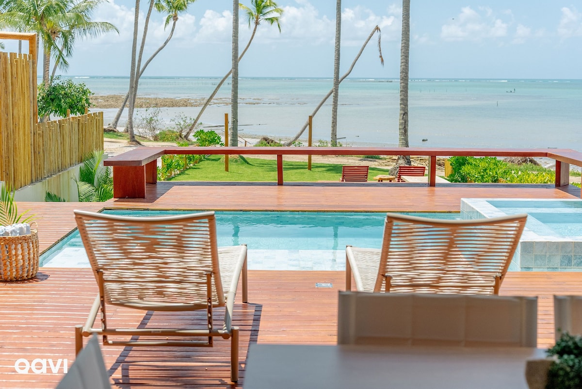 A wooden deck features two lounge chairs facing a tranquil ocean view. A salt-water pool is positioned nearby, and lush greenery surrounds the area. Distinctive palm trees provide a tropical atmosphere against a clear blue sky.