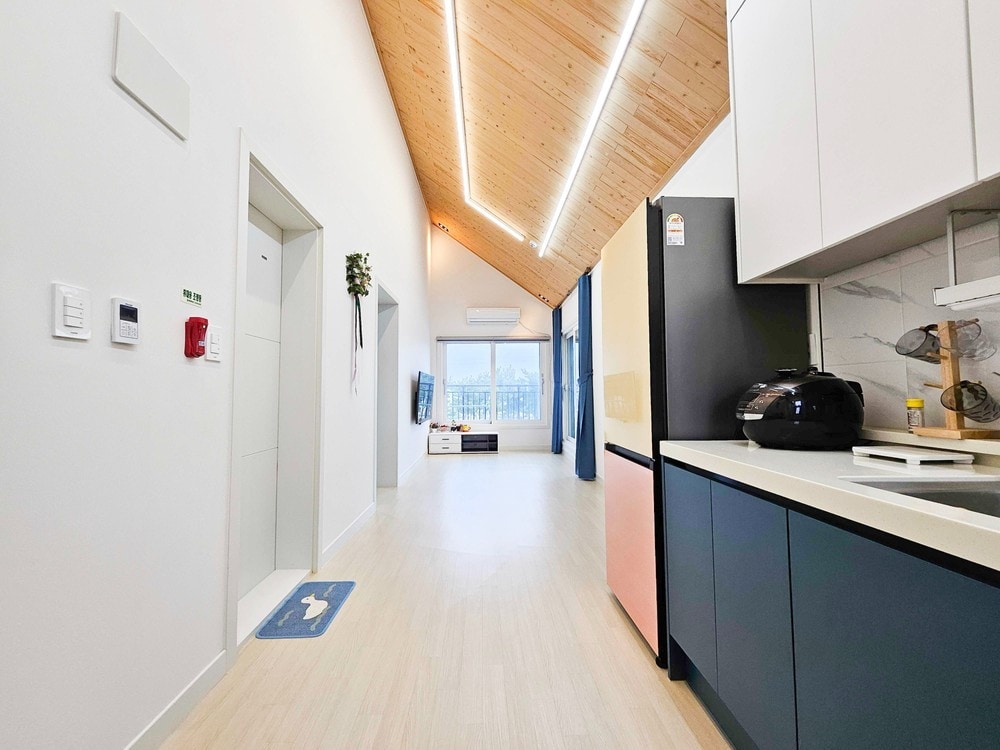 The living space features a high wooden ceiling, complemented by soft lighting along the beams. A kitchenette is visible on one side, with modern appliances, and a large window provides a view of the outdoor scenery, allowing natural light to fill the room.