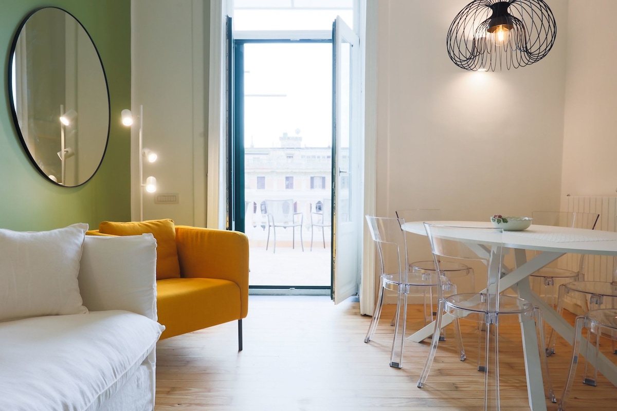 A bright living space is highlighted by an orange sofa and a round mirror on the wall. A glass dining table is surrounded by clear chairs, with doors opening to a terrace outside offering scenic views. Natural light filters in through large windows.