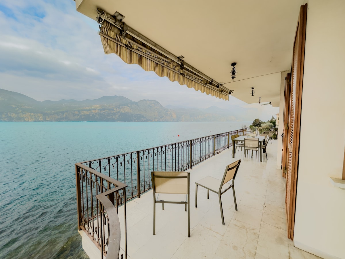 A balcony overlooking the serene lake features four chairs and a table, ideal for enjoying the view of the water and mountains. The space is framed by a decorative railing and an awning, providing shade while enhancing the lakeside atmosphere.