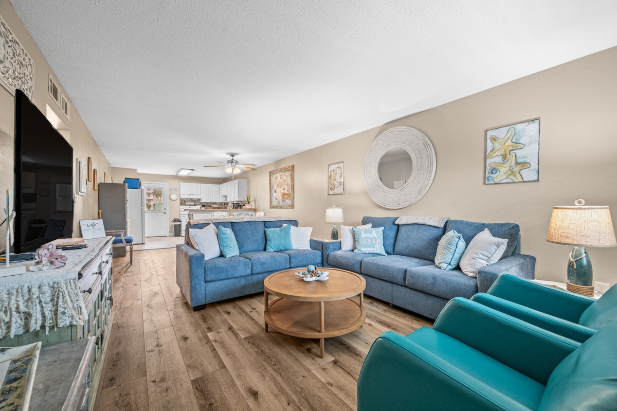A spacious living area features plush couches in shades of blue and a circular wooden coffee table at the center. Neutral walls are adorned with coastal-themed artwork, and a soft light illuminates the room from a nearby lamp. Open layout allows views of the kitchen in the background.