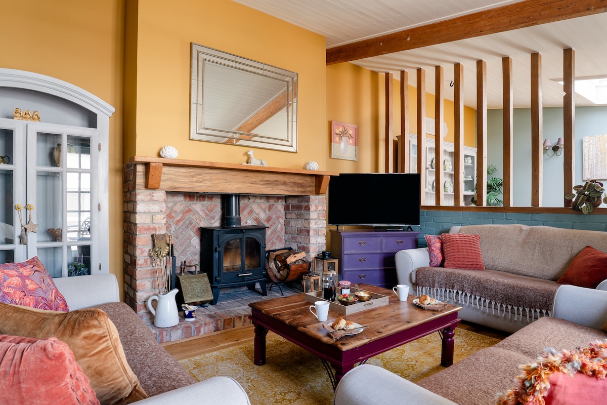 The inviting living area features a cozy wood burner set within a brick fireplace. Two sofas are adorned with colorful cushions, arranged around a wooden coffee table that holds breakfast items. A large mirror above the mantel reflects warm tones from the painted walls.