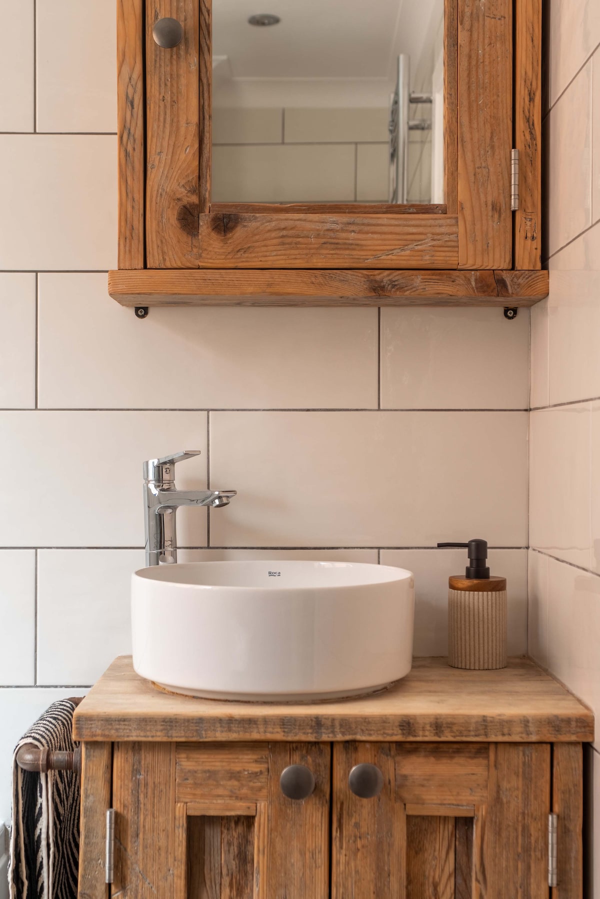 A rustic bathroom sink area features a modern round basin atop a wooden cabinet, complemented by a discreet pump bottle. A mirrored cabinet is mounted above for additional storage and convenience. The tiles are laid in a clean, grid pattern that enhances the overall design.