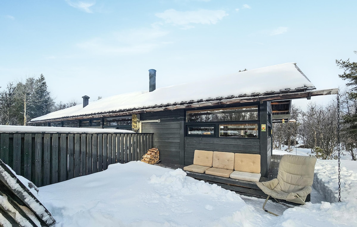 A simple cabin exterior is showcased, surrounded by fresh white snow. The dark wooden structure features large windows and a covered porch with comfortable seating. Firewood is stacked nearby, and trees can be seen in the background, creating a serene winter landscape.