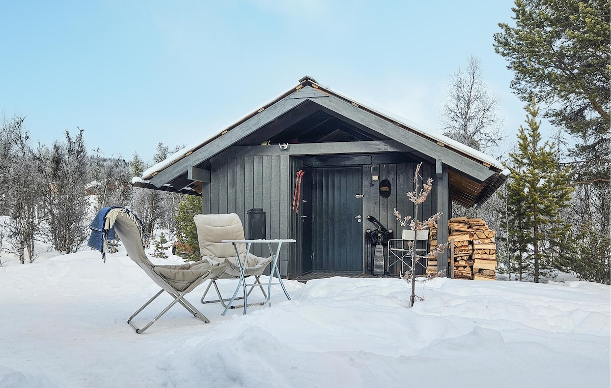 A charming cabin is set against a snowy landscape. In front, a small table and two chairs are positioned on a white expanse. Firewood is stacked neatly beside the entrance, and trees are visible in the background beneath a clear sky.