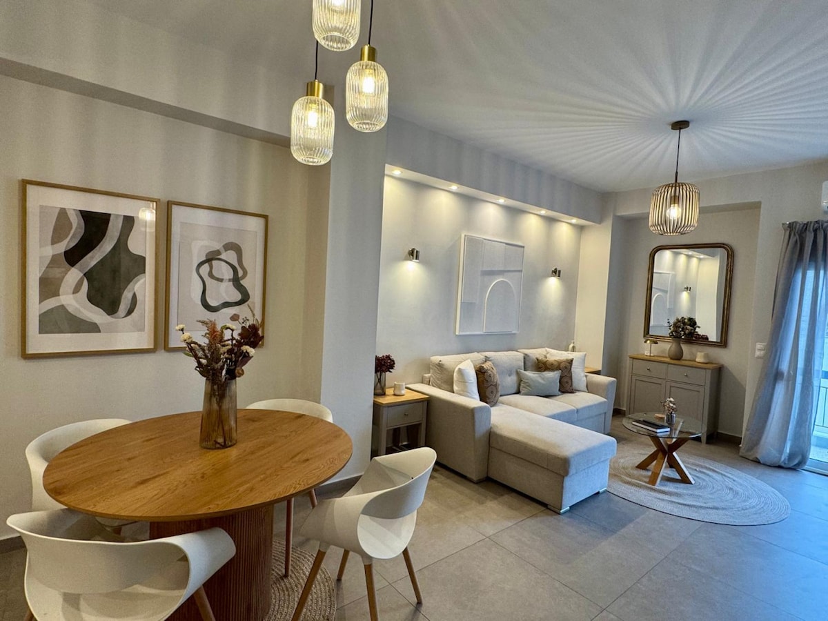 A contemporary living space is highlighted, featuring a round wooden dining table accompanied by four white chairs. A comfortable sectional couch is arranged in the lounge area, with soft decorative pillows. Wall art and modern lighting fixtures enhance the ambiance within the room.