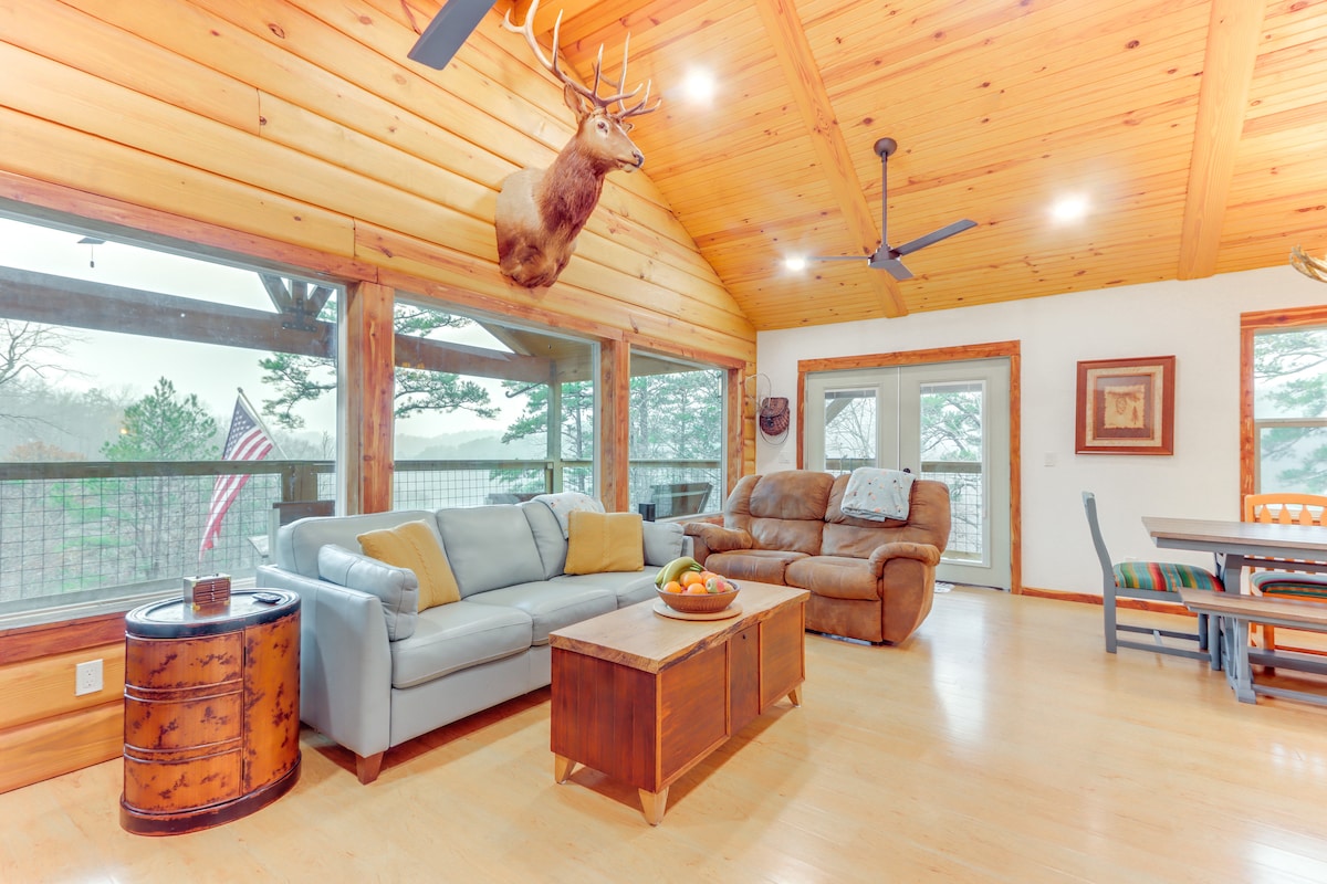 A spacious living area features wooden walls and high ceilings, complemented by large windows that invite natural light. A comfortable sofa and two armchairs provide seating. A rustic coffee table sits in the center, while a dining table and chairs are visible in the background.