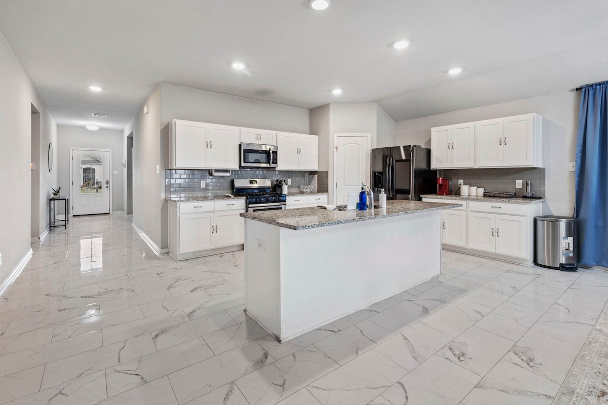 A spacious kitchen features modern white cabinetry and granite countertops. Stainless steel appliances are positioned for convenient access, while the open layout allows for easy movement. Recessed lighting illuminates the area, complemented by a large window dressed with a blue curtain, providing natural light.