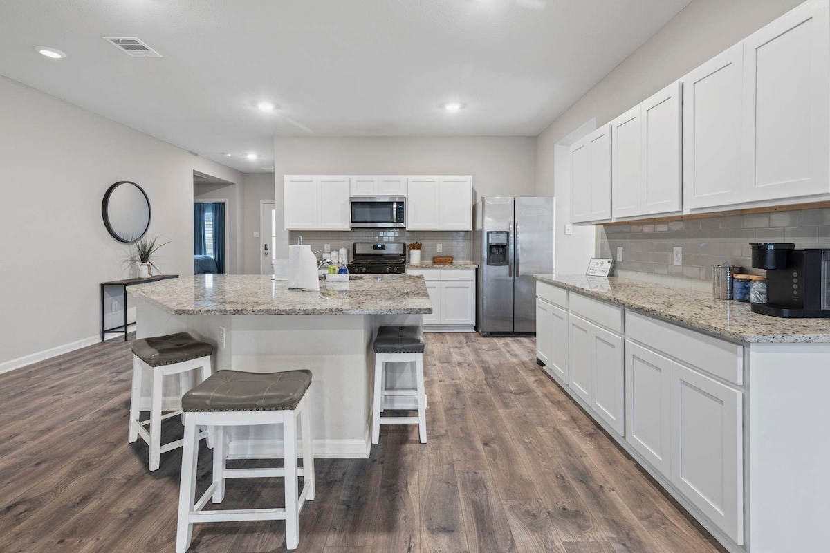 A spacious kitchen is shown with modern white cabinetry and granite countertops. The kitchen island features two stools, providing a casual dining option. Stainless steel appliances are neatly arranged, and a dining area is partially visible in the background, enhancing the home’s open layout.