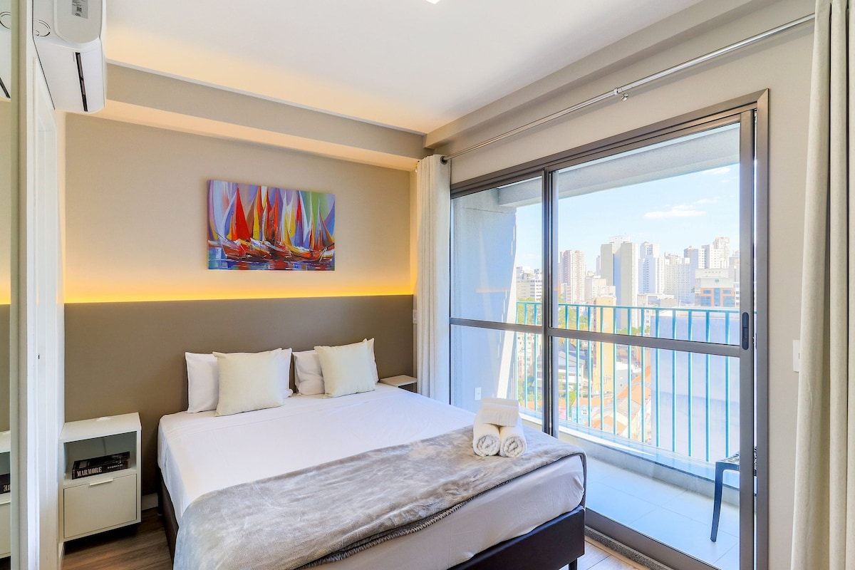 A bright and modern sleeping area features a spacious bed with fresh linens, accompanied by two decorative pillows. A colorful artwork hangs above the bed, and large glass doors lead to a balcony offering city views. Natural light fills the room, accentuating its contemporary design.