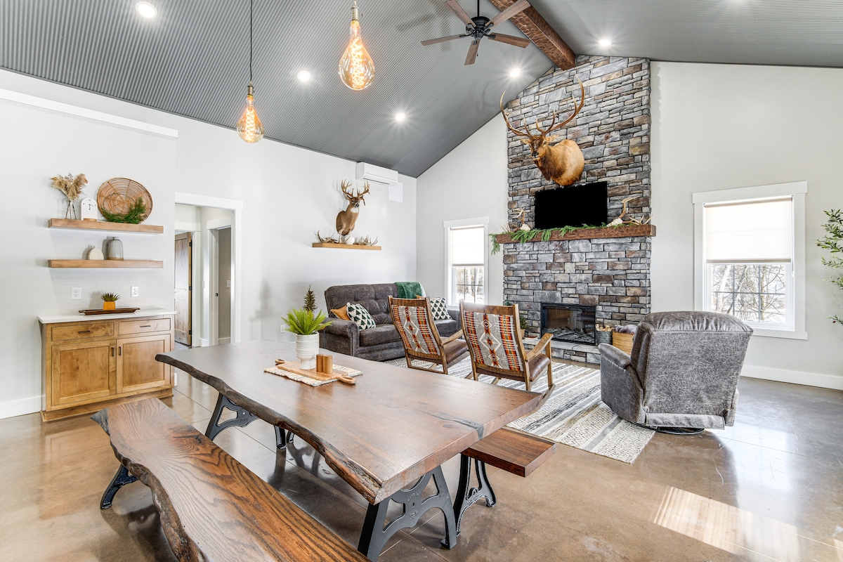 A spacious living area integrates a wooden dining table with a natural finish and seating for six. A stone fireplace serves as a focal point, accompanied by taxidermy decor. Comfortable seating options, including two armchairs and a sofa, create an inviting space illuminated by pendant lights.