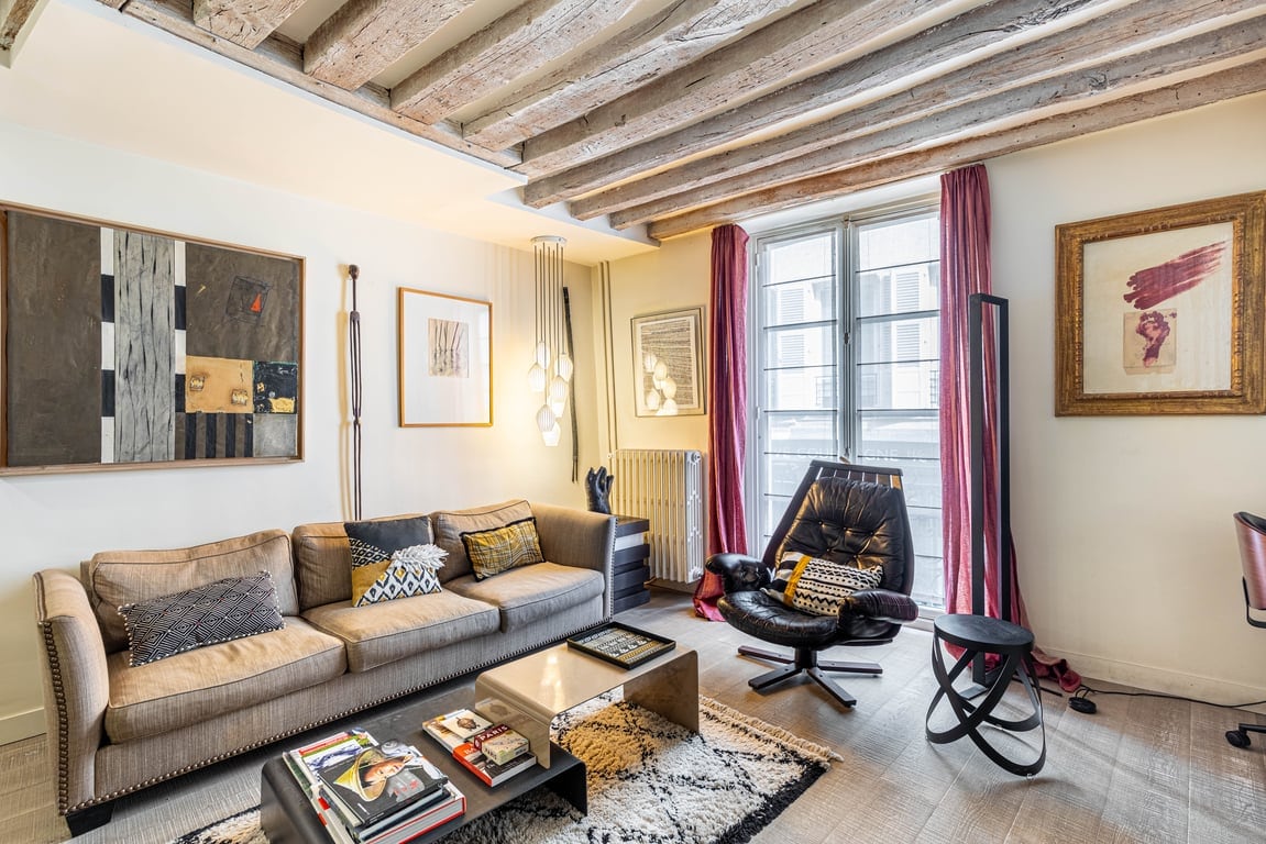 A living area features a comfortable sofa with textured cushions and a glass coffee table. Art is displayed on the walls, and light fixtures hang from the ceiling. The space is illuminated by natural light from large windows covered by soft drapes.