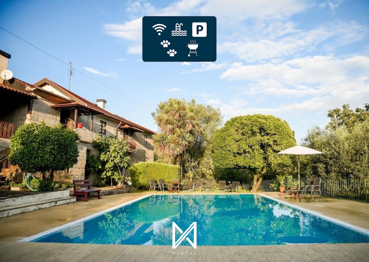 A well-maintained private pool is situated in the foreground, surrounded by a spacious outdoor area. Lush greenery and trees frame the property, while seating and sunshade provide inviting spots for relaxation. The two-story house, styled with natural stone, enhances the tranquil ambiance.