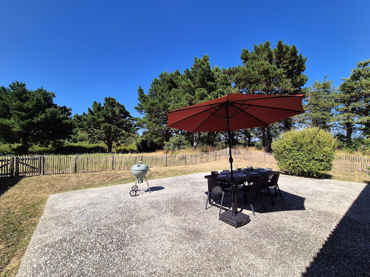 A spacious outdoor area features a large umbrella providing shade over a table with chairs. A barbecue grill is situated nearby, surrounded by greenery and a fenced perimeter, creating a serene environment perfect for relaxation.
