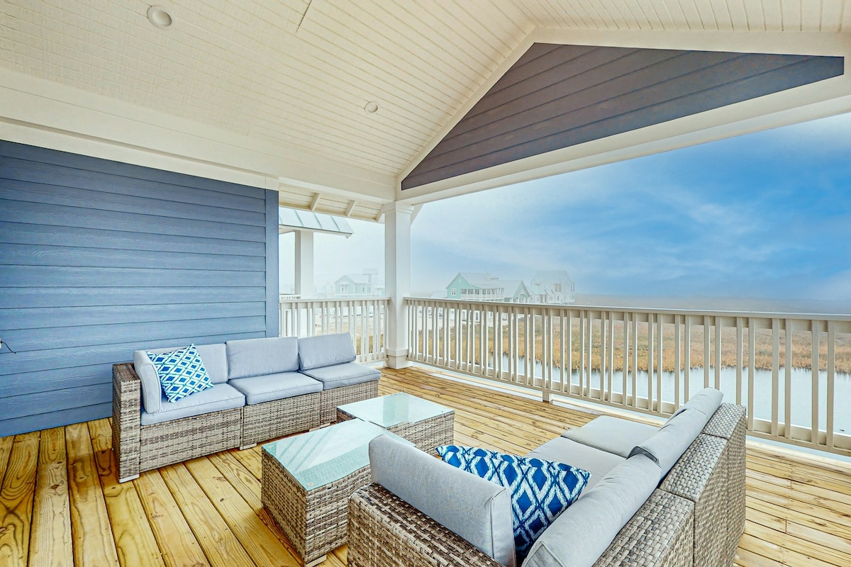 A covered outdoor seating area features a light-colored sectional sofa and a glass-top coffee table. The view includes a serene landscape beyond a railing, with soft blue skies and hints of water visible.