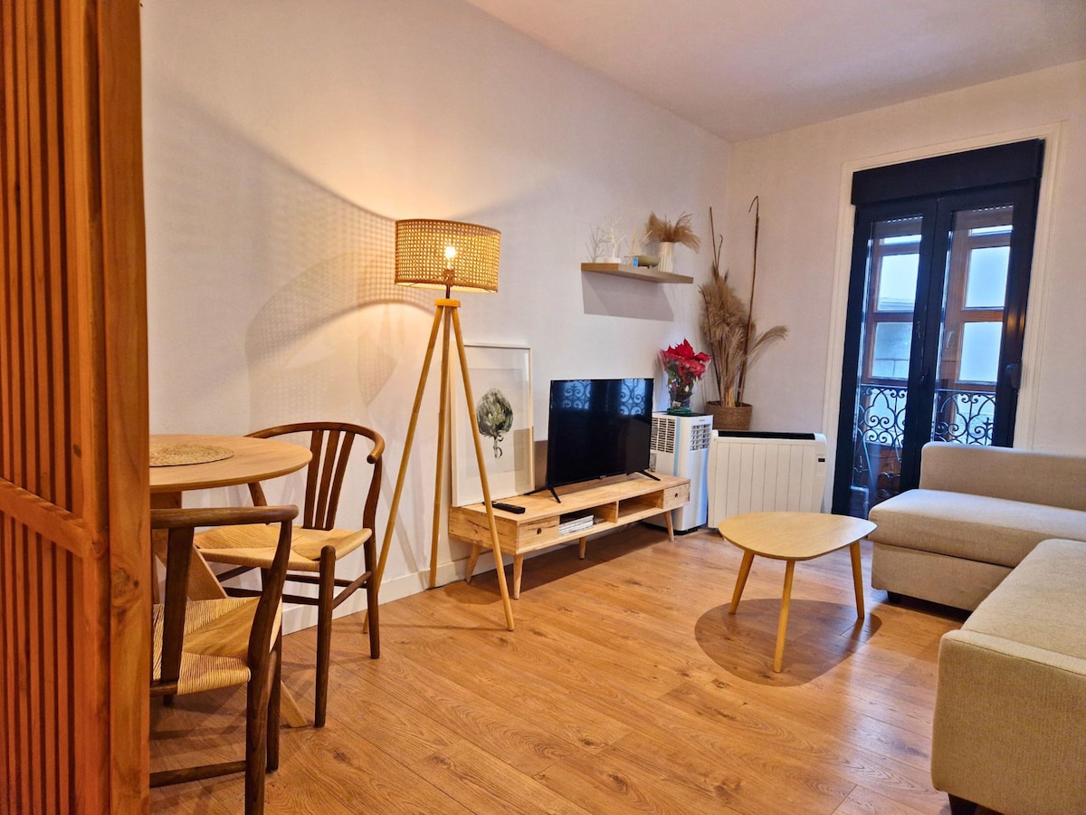 A cozy living-dining area is equipped with a small round table and two wooden chairs. A light fixture provides gentle illumination beside the TV, which is placed on a low wooden shelf. The sofa and light wooden coffee table enhance the space's welcoming ambiance.