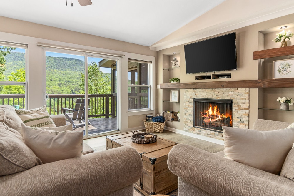 A spacious living room features a comfortable seating area with light-colored sofas arranged around a rustic coffee table. A stone fireplace adds a cozy touch, while large sliding glass doors open to a deck, offering views of lush greenery and distant mountains.
