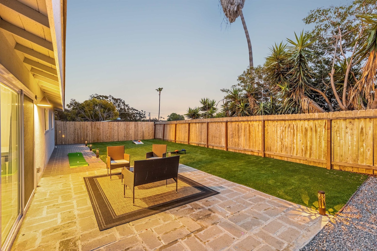 A spacious yard is presented, featuring a well-manicured lawn and a stone patio. Comfortable seating is arranged around a small rug, and a wooden fence offers privacy. Lush palm trees and greenery add a natural touch, creating an inviting outdoor space.