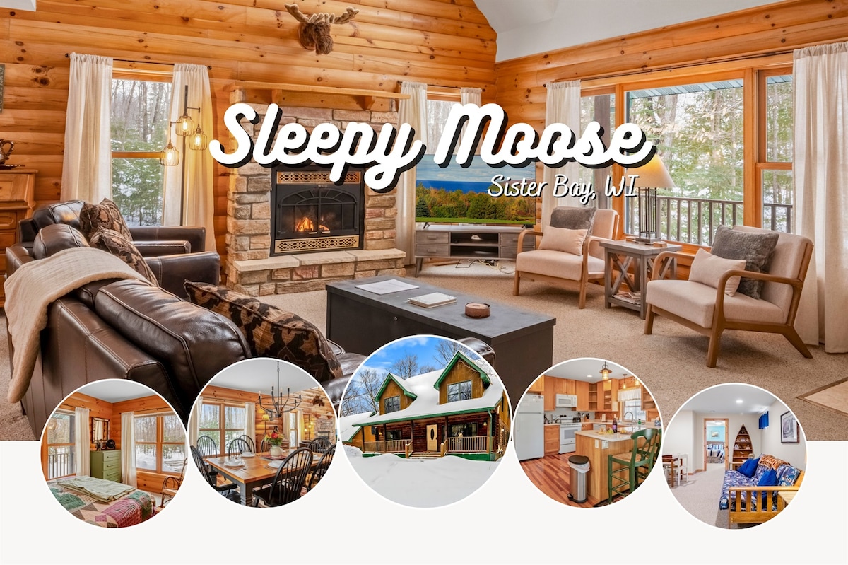 The spacious living room features a large stone fireplace surrounded by wooden accents and comfortable seating. Ample natural light is provided by large windows, creating a welcoming ambiance. A flat-screen TV is positioned across from the seating area, enhancing the cozy atmosphere.