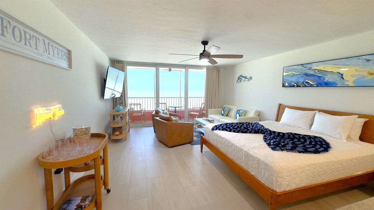 A spacious room features a large bed with crisp white linens, accompanied by a decorative blue throw. A seating area with a leather chair and a mounted flat-screen TV is visible. Expansive glass doors provide natural light and views of the balcony overlooking the beach.