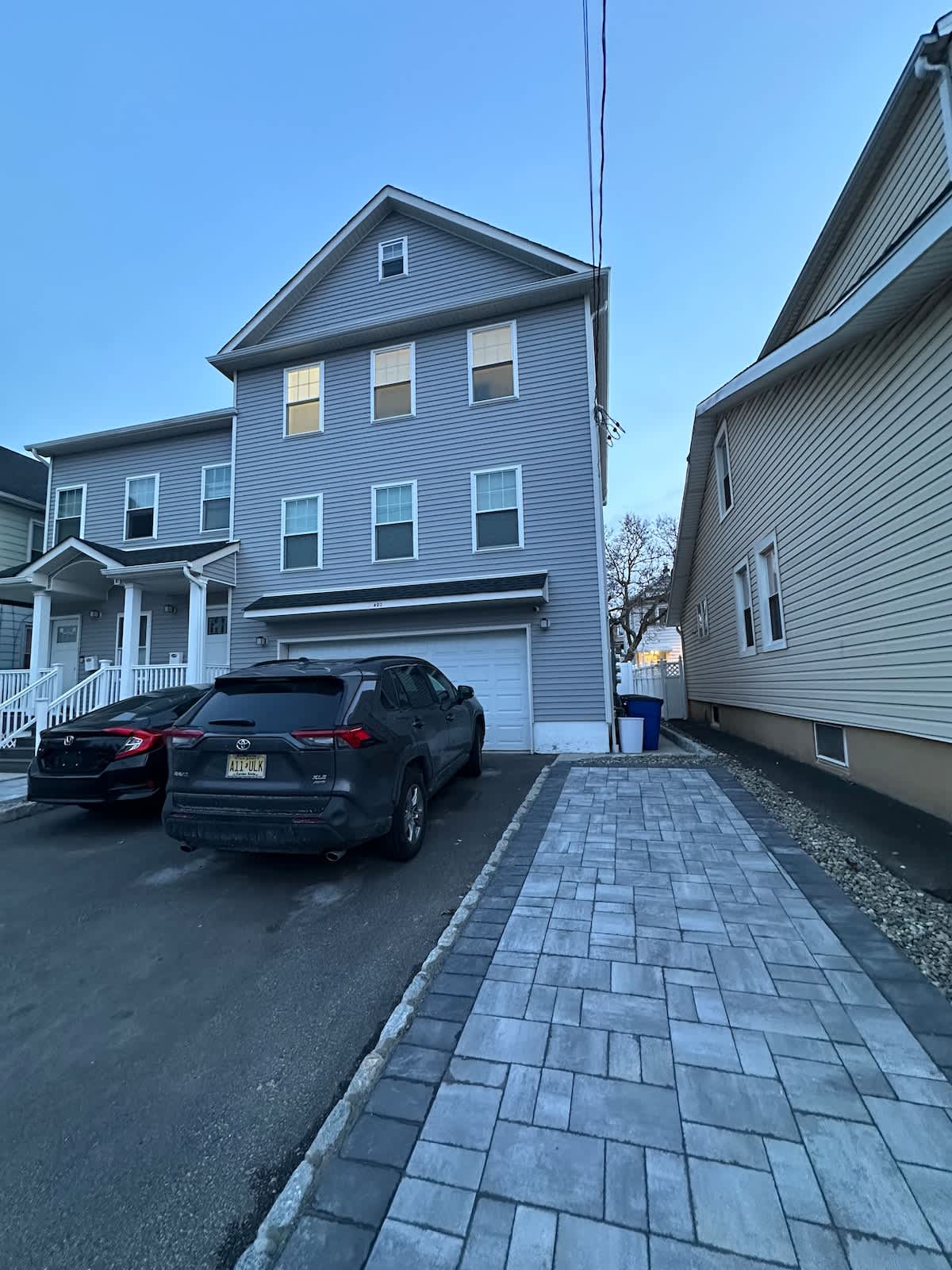 The exterior of a multi-story building is shown, featuring a light grey façade. A driveway accommodates two parked vehicles, with a sidewalk made of stone pavers leading alongside. The setting is enhanced by adjacent buildings that reflect a residential neighborhood.