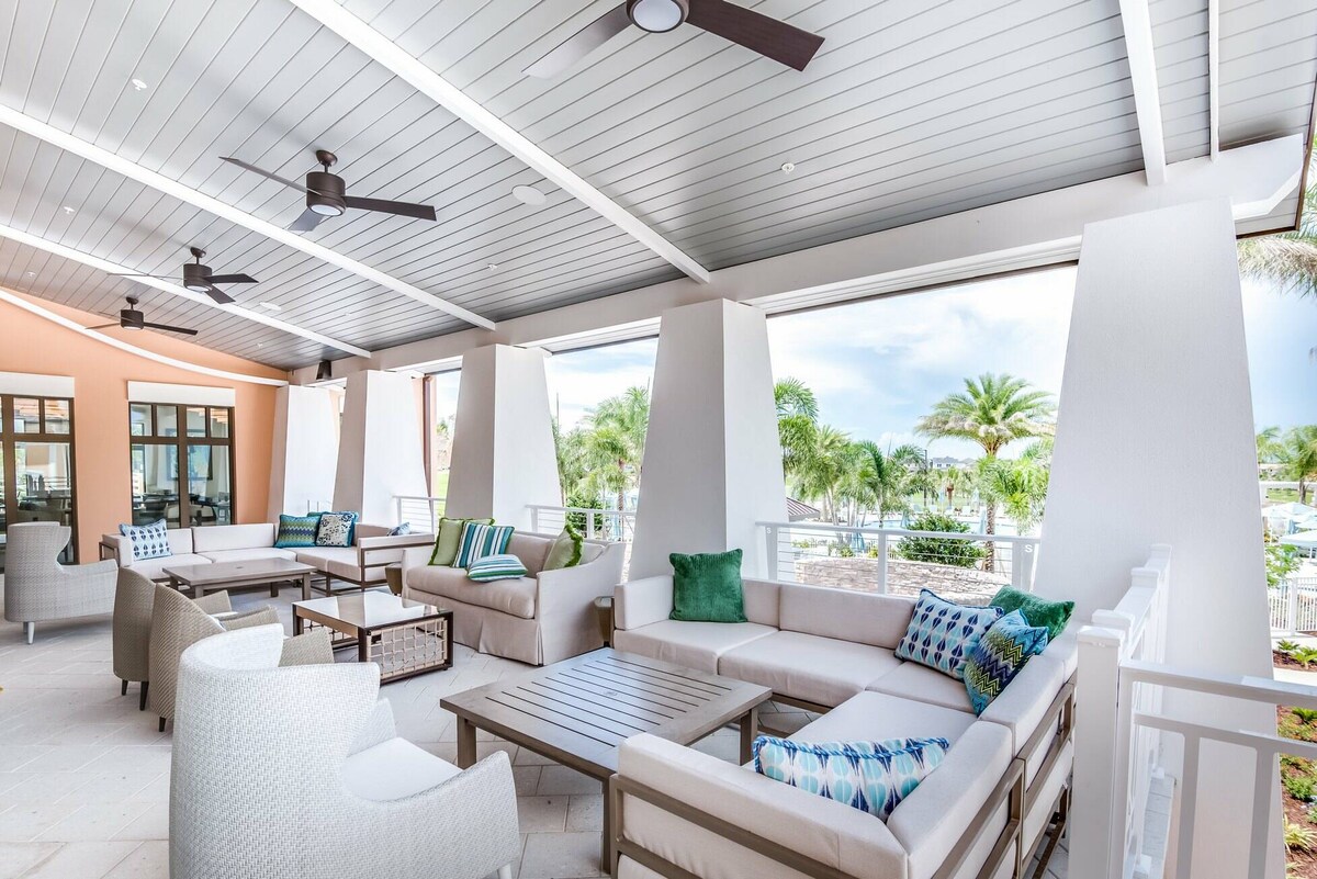 An outdoor lounge area features modern seating arrangements with plush cushions in soft colors. Large windows span the space, offering views of palm trees and greenery. Ceiling fans provide comfort, while a stylish coffee table sits at the center, enhancing the inviting atmosphere for relaxation.
